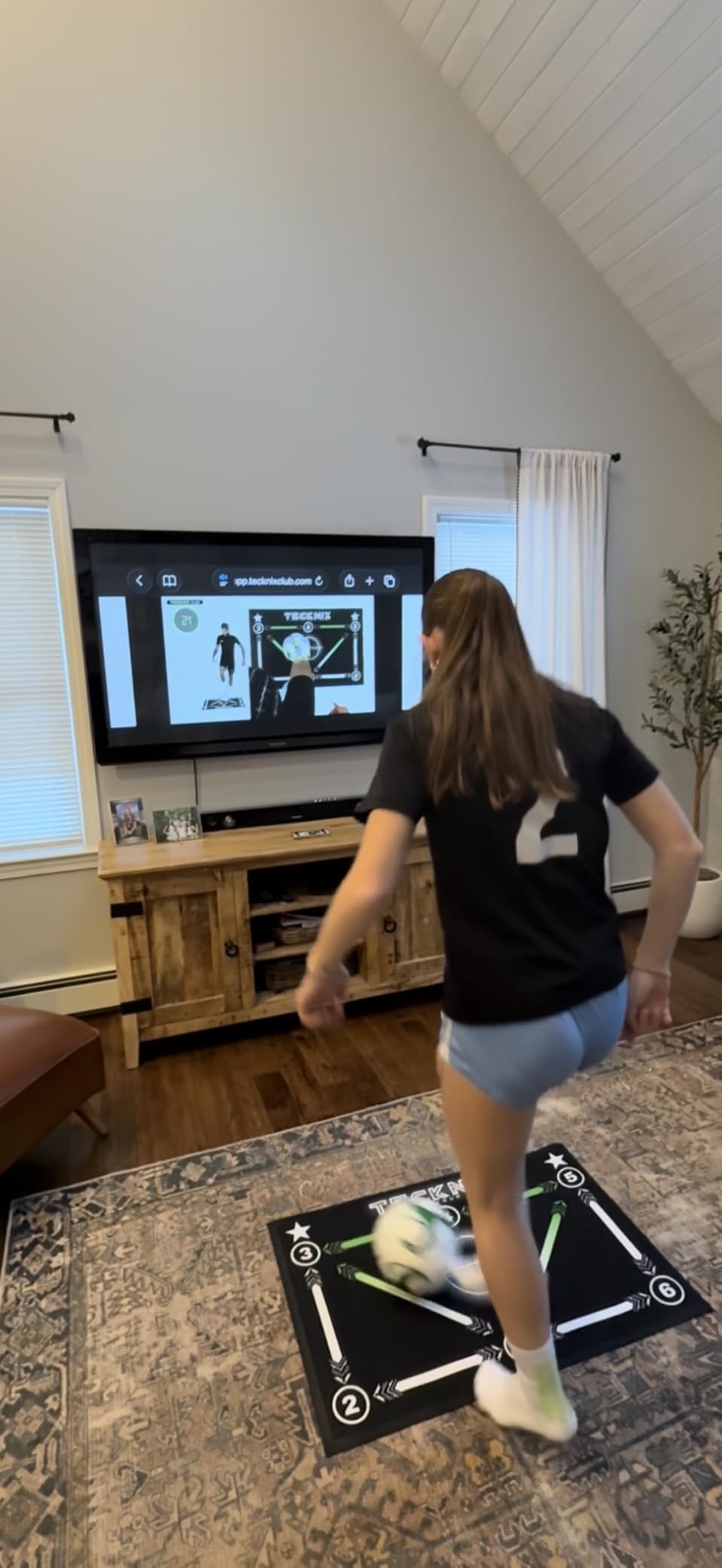 Girl in a soccer jersey practicing on a training mat in a living room while a training video plays on the TV