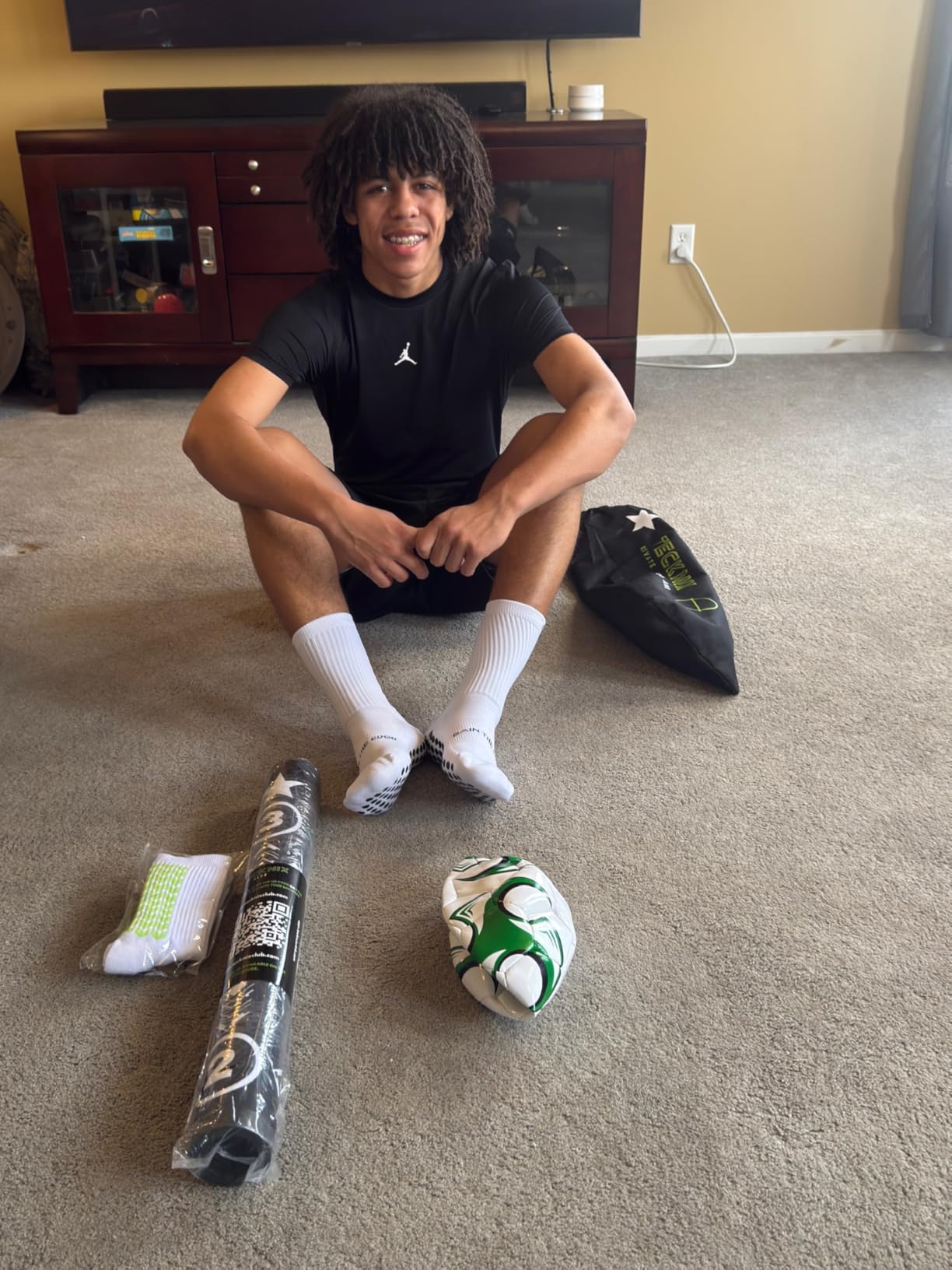 Teenage player in grip socks sitting on a carpet next to a rolled-up training mat, a ball and a drawstring bag